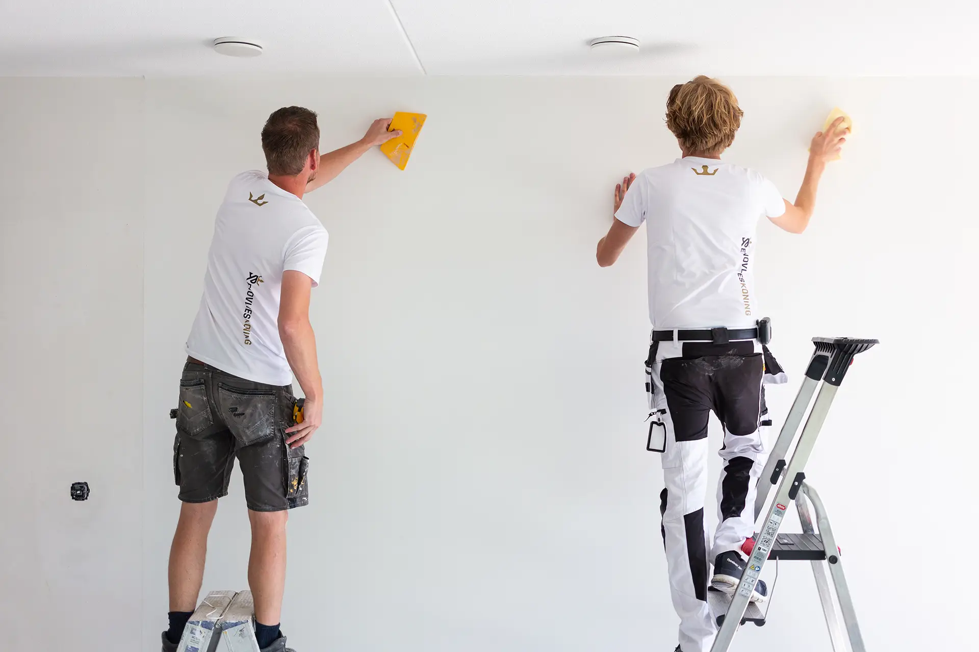 Twee professionele renovlies behangers in actie. Een behanger staat op een trap terwijl de ander op de grond werkt. Beiden gebruiken gereedschap om een muur nauwkeurig voor te bereiden voor renovliesbehang.