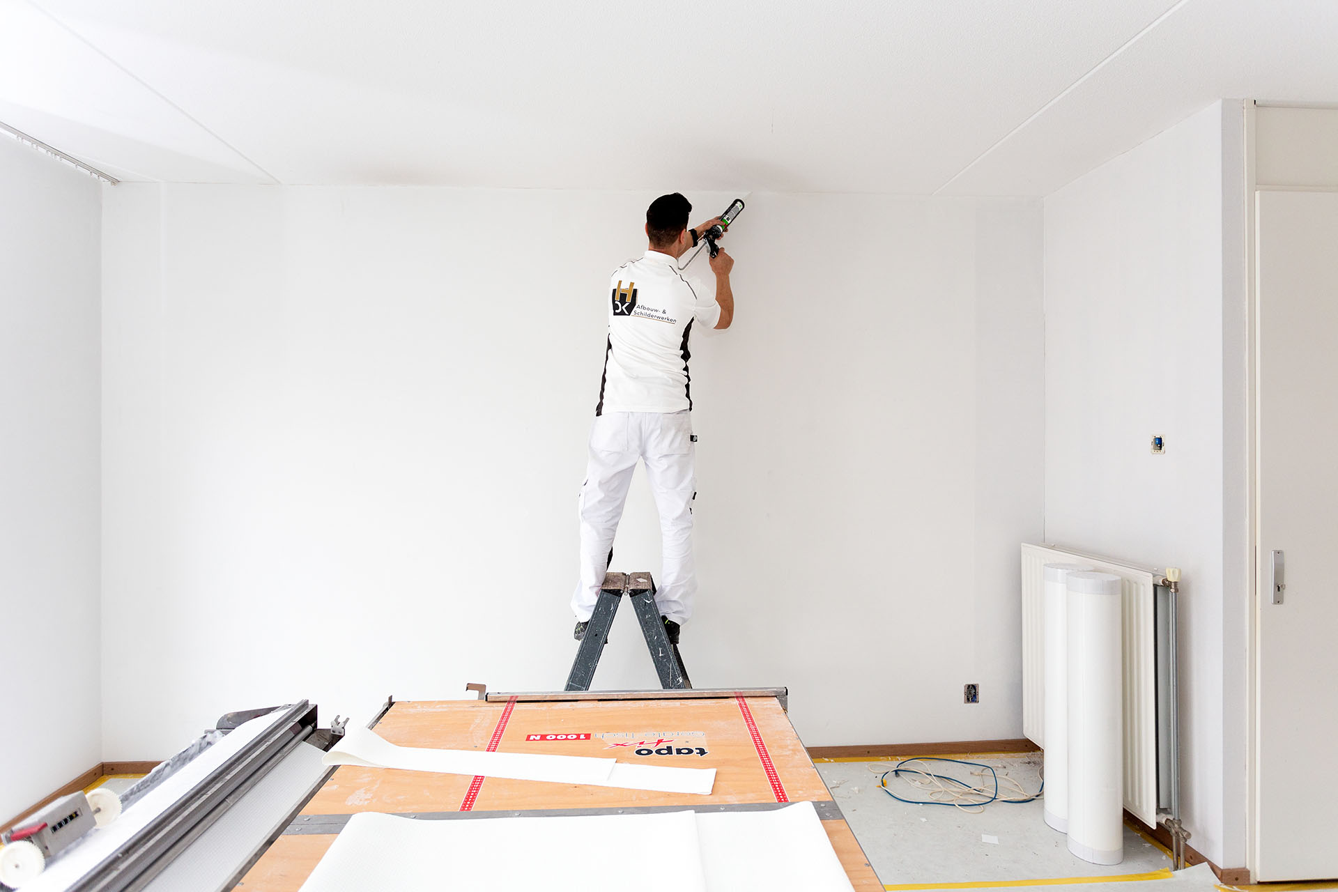 Een schilder staat op een ladder in een lege kamer en brengt kit aan op de bovenkant van een muur. Op de vloer liggen gereedschappen en materialen