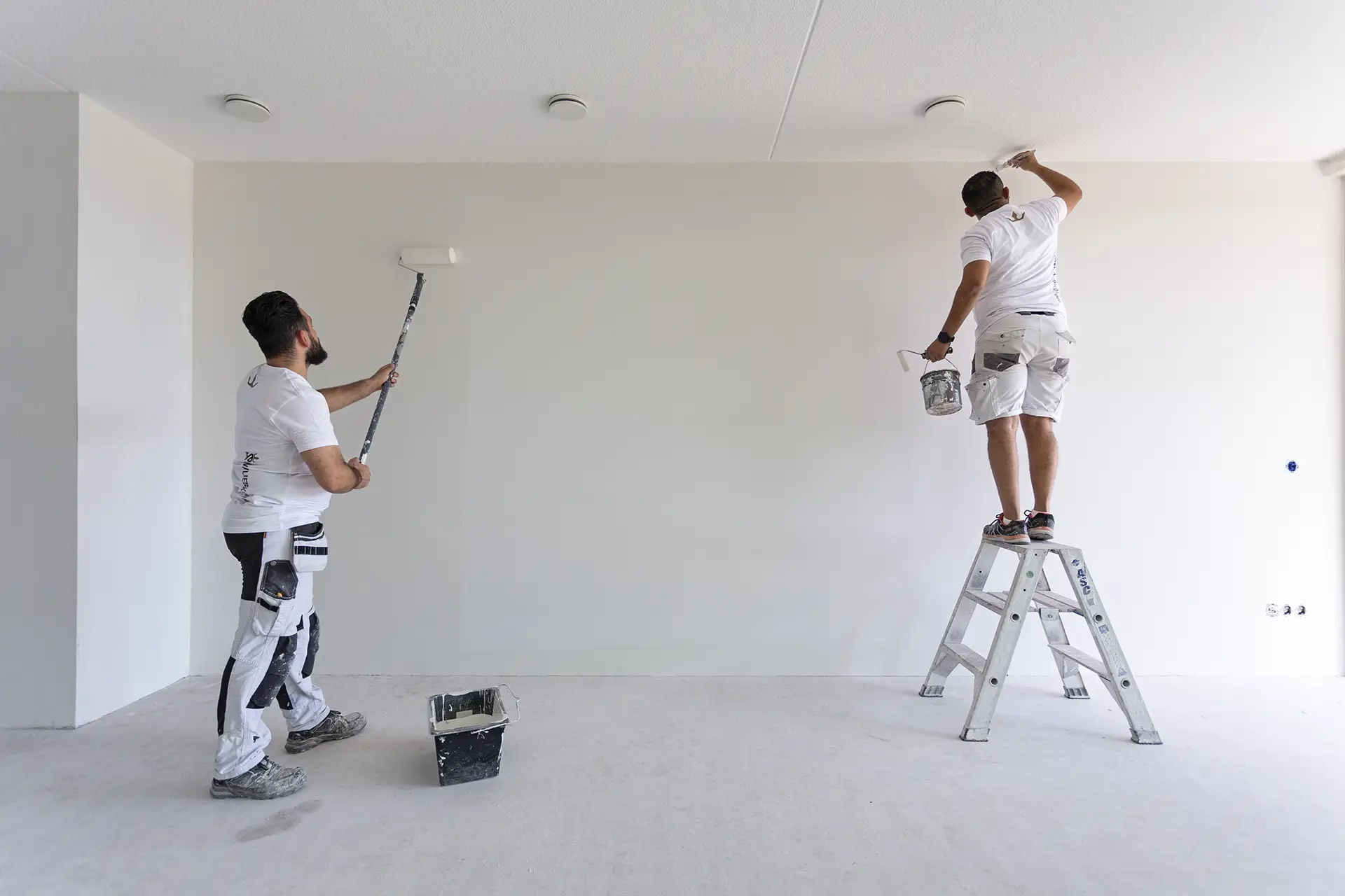 Vakmannen Veenendaal bezig met het schilderen van het renovlies. Eén behanger werkt met een verfroller, terwijl de ander de randen bijwerkt.