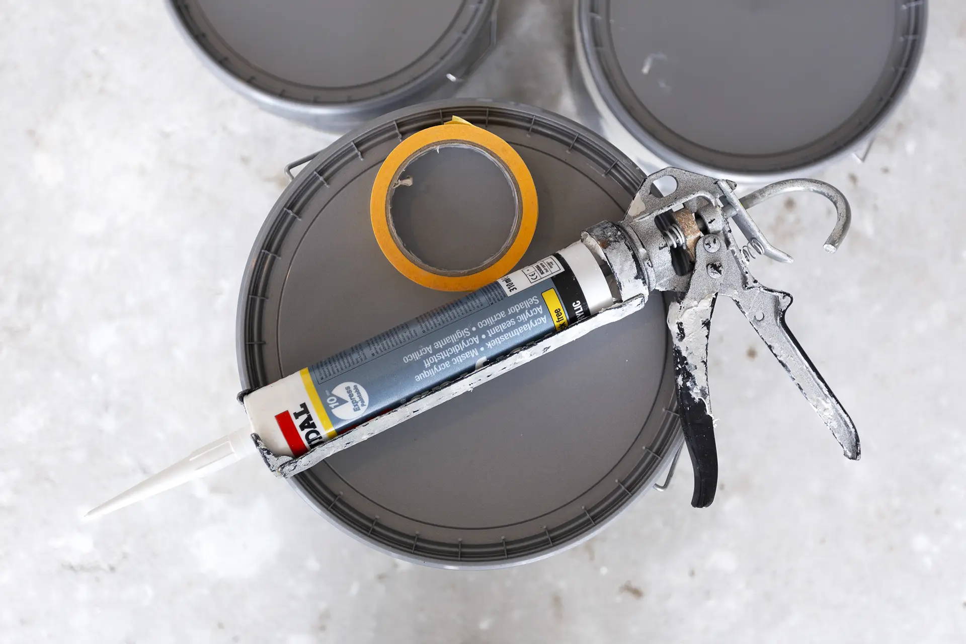 Een kitpistool met een tube afdichtmiddel ligt bovenop een grijze emmer, omringd door een rol geel afplaktape en extra emmers op de achtergrond. Ideale benodigdheden voor een professioneel renovatieproject in Amersfoort