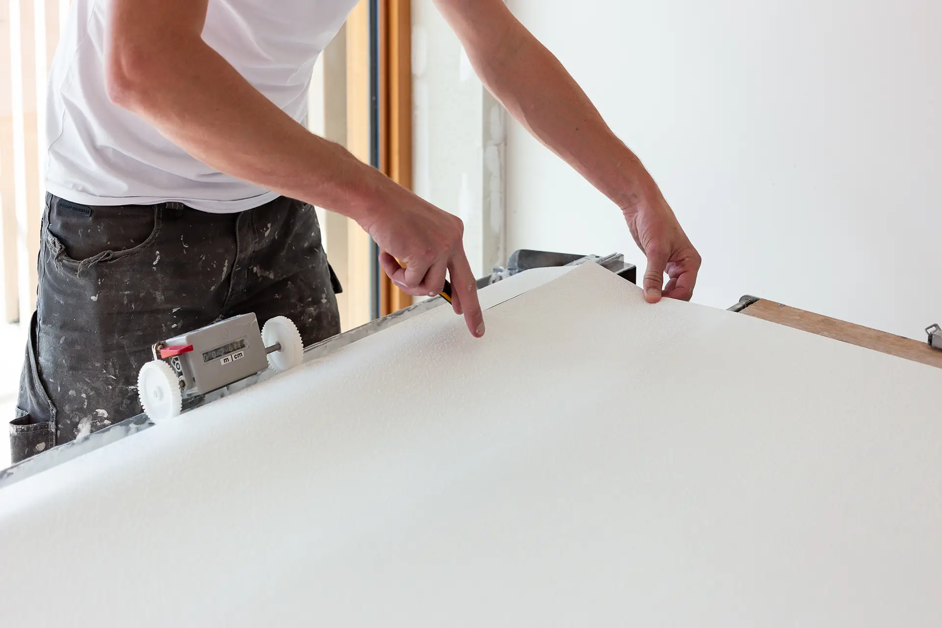 Een persoon meet en snijdt renovlies nauwkeurig op een plat oppervlak. De persoon draagt een witte trui en zwarte verfbroek, terwijl gereedschap en materiaal klaar liggen voor een professionele wandafwerking in Barneveld